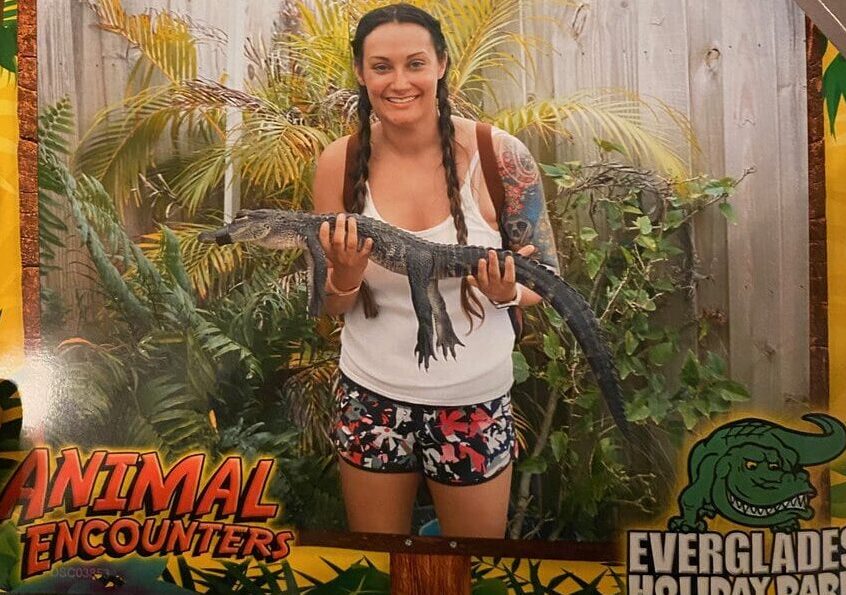 A smiling woman with braided hair holds a small alligator in front of tropical plants and a wooden fence, standing between signs that read Animal Encounters and Everglades Holiday Park.