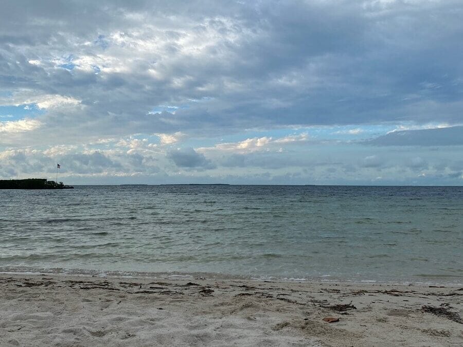 A sandy beach with gentle waves under a partly cloudy sky; the horizon separates the calm sea from the blue and gray clouds, with a small patch of land visible to the left.