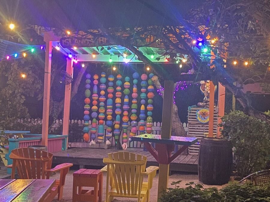A cozy outdoor patio with colorful Adirondack chairs, string lights, and a vibrant wall decorated with hanging painted buoys under a wooden canopy at night.