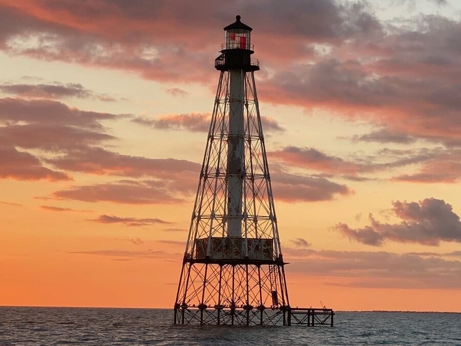 A tall, skeletal lighthouse stands in the ocean at sunset, silhouetted against an orange and pink sky with scattered clouds. The lighthouses red light is clearly visible at the top.
