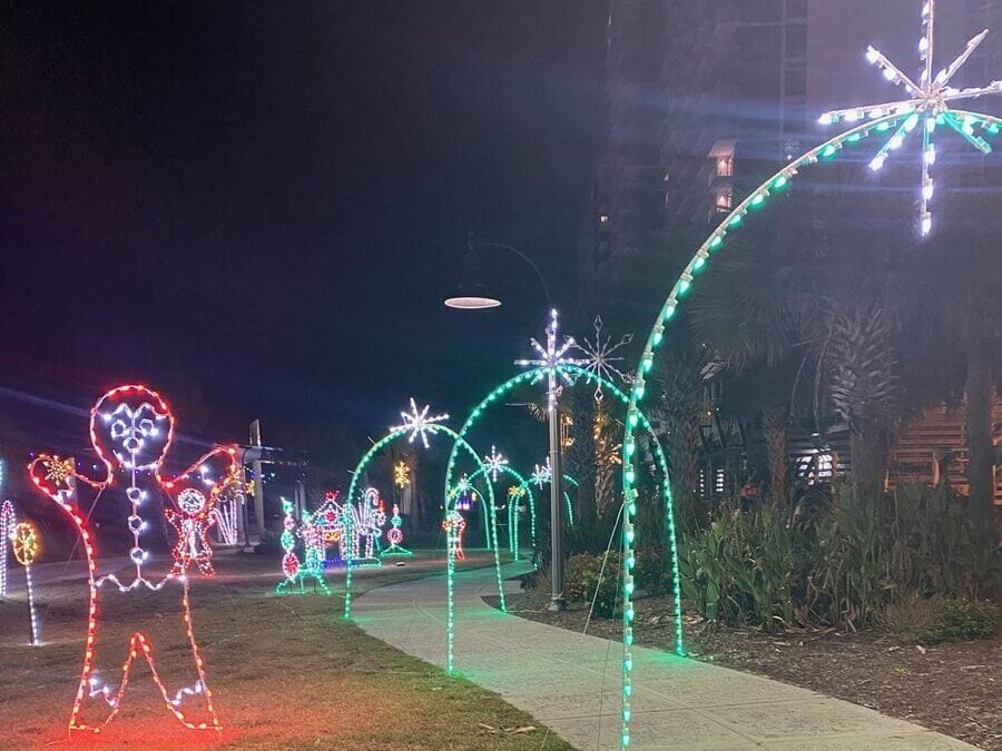 A nighttime outdoor scene shows festive holiday lights, including a large glowing gingerbread figure and illuminated arches with star-shaped tops, lining a sidewalk with greenery on the side.