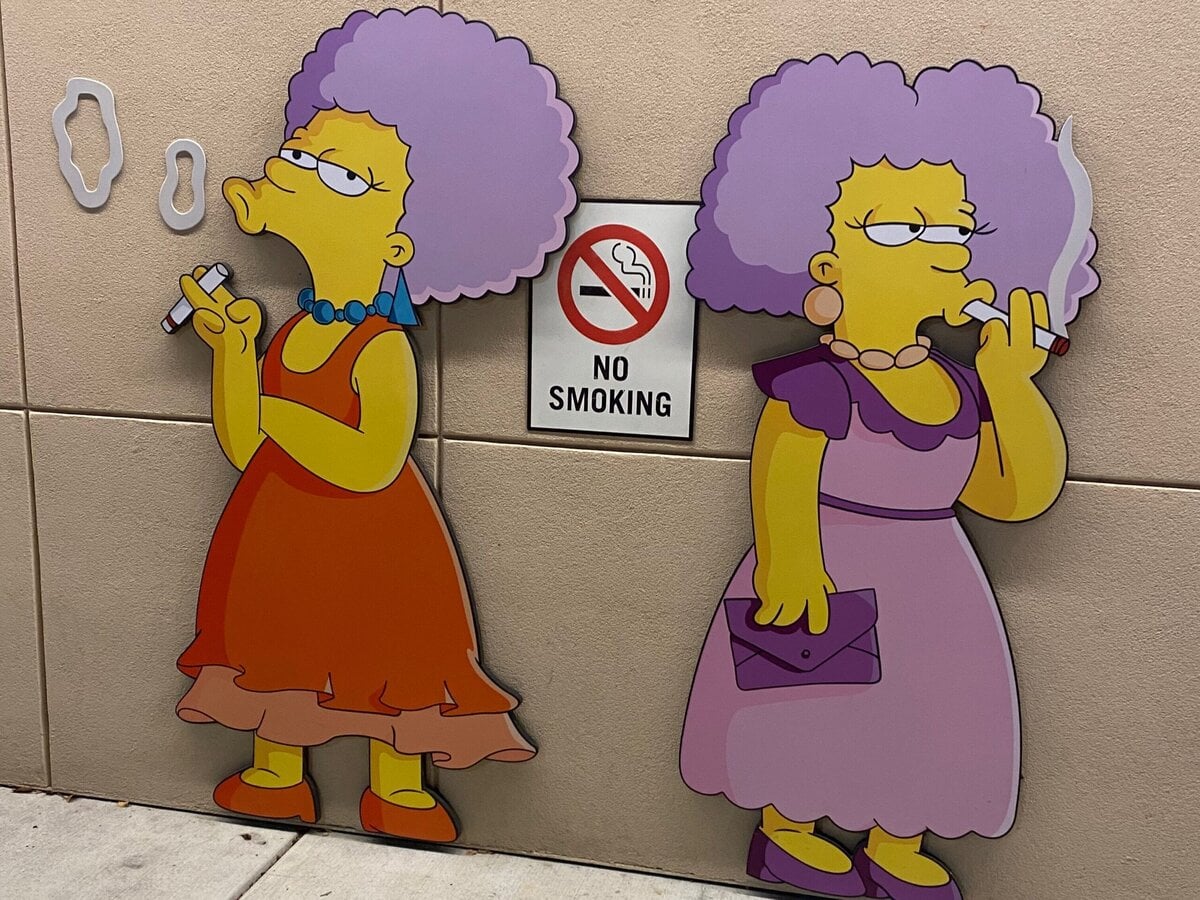 Two cartoon women with purple hair, identified as characters from The Simpsons, are smoking cigarettes while standing on either side of a No Smoking sign mounted on a beige wall.