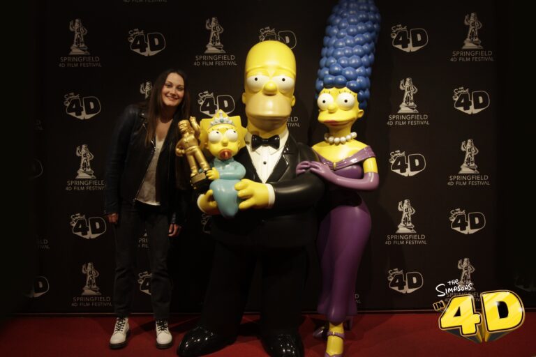 A woman smiles and poses next to life-sized statues of Maggie, Homer, and Marge Simpson in formal attire at a Simpsons in 4D event—one of the fun things to do alone in Myrtle Beach—with a branded backdrop behind them.