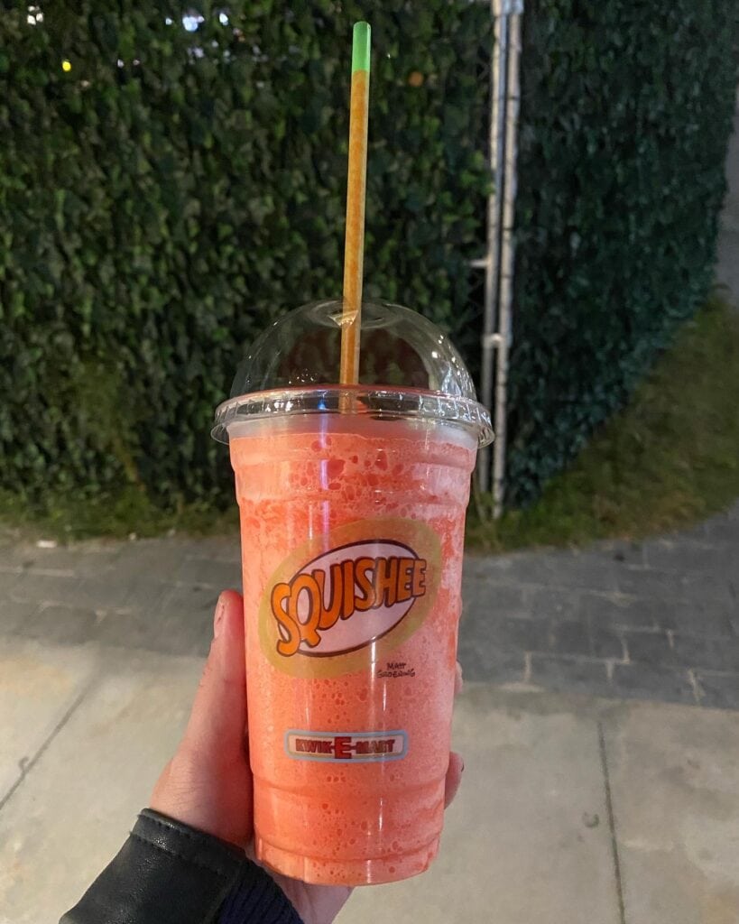 A hand holds a clear cup with a dome lid and straw, filled with a bright orange frozen Squishee—perfect for sipping while exploring nighttime greenery and sidewalks, one of the fun things to do alone in Myrtle Beach.