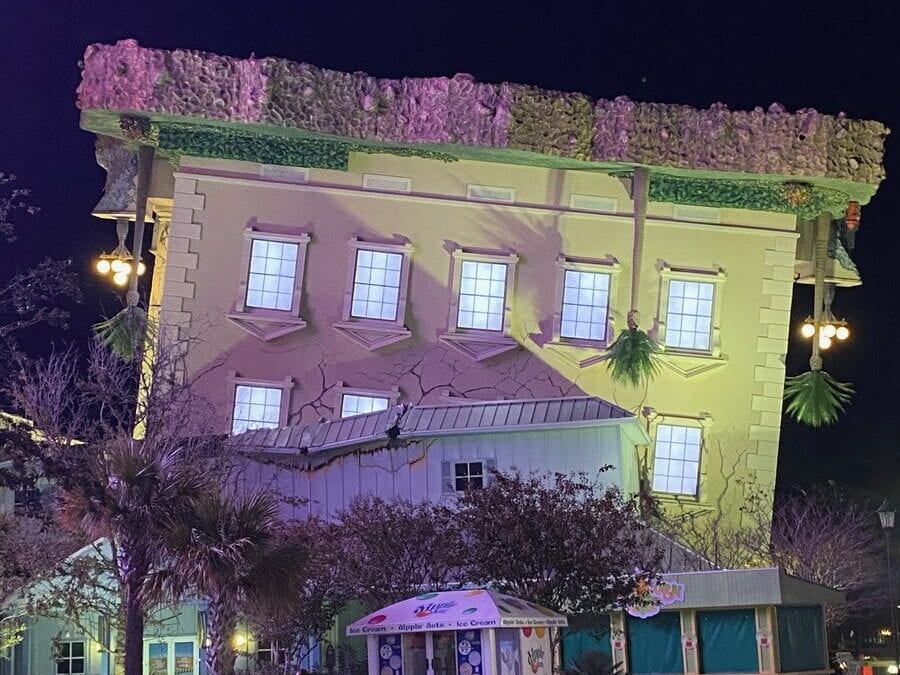 A building designed to appear upside down, with its roof on the ground and windows and lamps inverted, illuminated by colorful lights at night. Trees and an ice cream stand are in front.