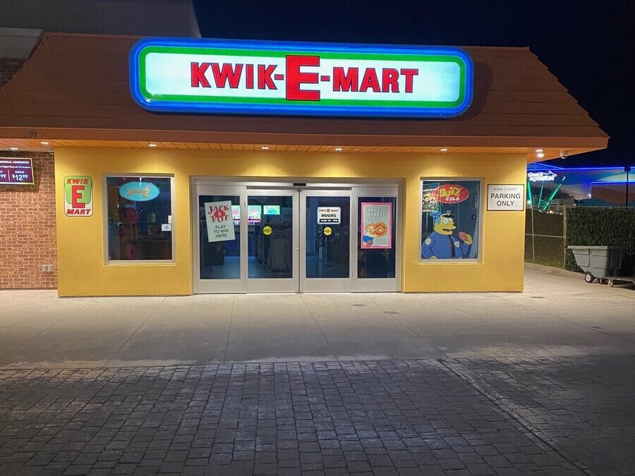 A brightly lit storefront at night displays a sign reading Kwik-E-Mart above double glass doors, with colorful cartoon posters in the windows and a Parking Only sign to the right.