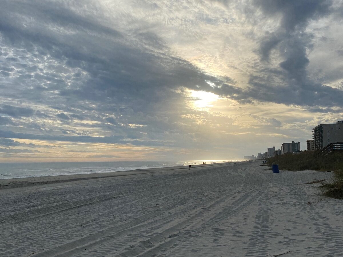 A sandy beach at sunrise or sunset with tire tracks in the sand, a few distant people walking, ocean waves, buildings along the right, and a dramatic cloudy sky with sunlight breaking through.