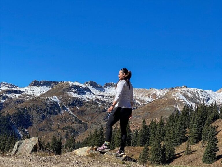 A woman in athletic clothing stands on a rocky path overlooking snow-capped mountains and pine trees under a clear blue sky.