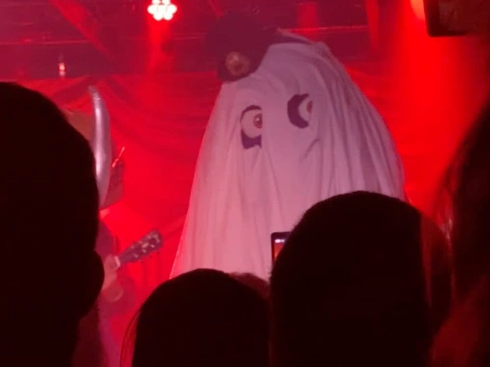 A person in a ghost costume with painted eyes and a cap stands on stage under red lighting, while another person in a horned helmet plays guitar. Audience silhouettes are visible in the foreground.