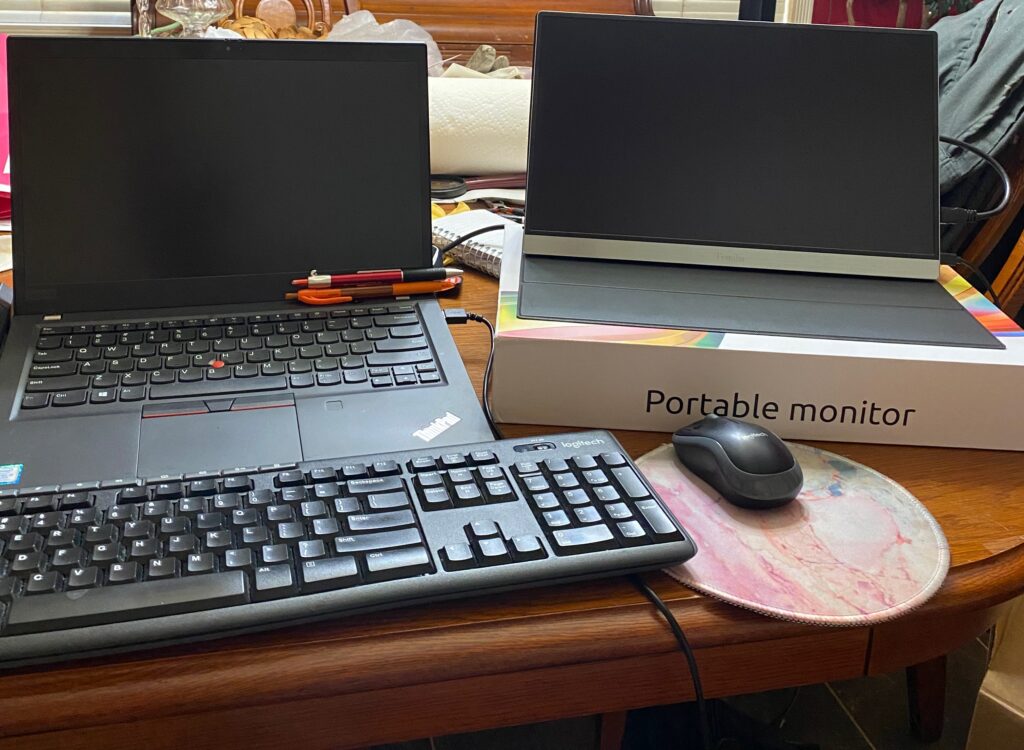 A laptop with pens on the keyboard sits next to a portable monitor on its box. A Logitech keyboard and mouse on a pink mouse pad complete this setup of digital nomad essentials, all arranged neatly on a wooden table.