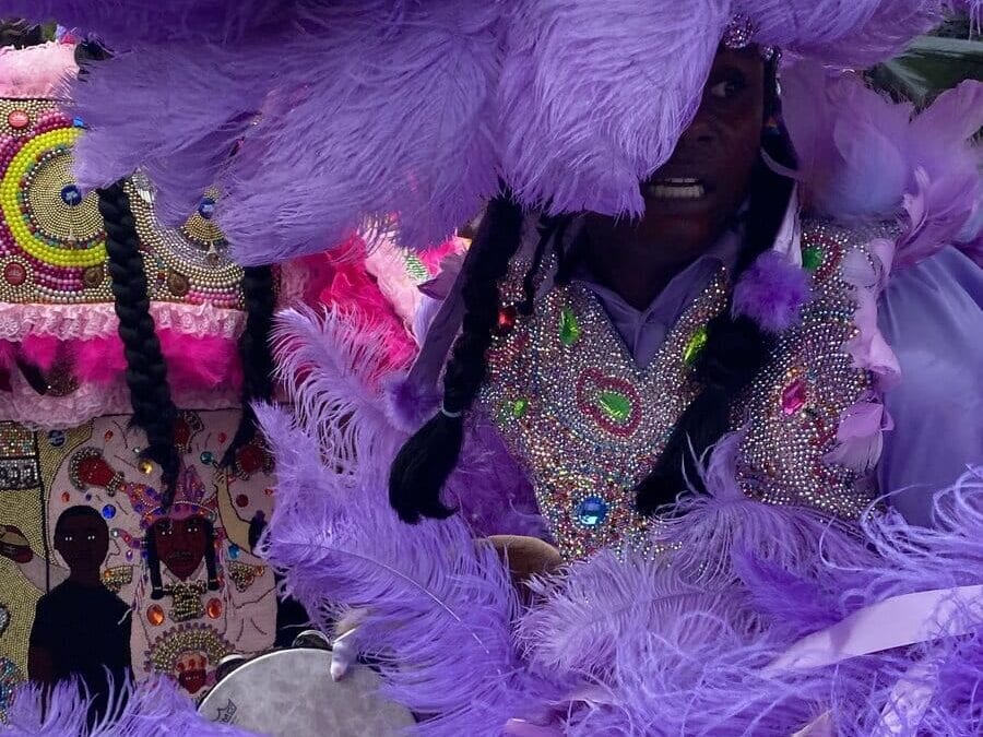 A person dressed in a vibrant costume adorned with beaded designs, sequins, and large purple feathers participates in a festive parade or celebration.