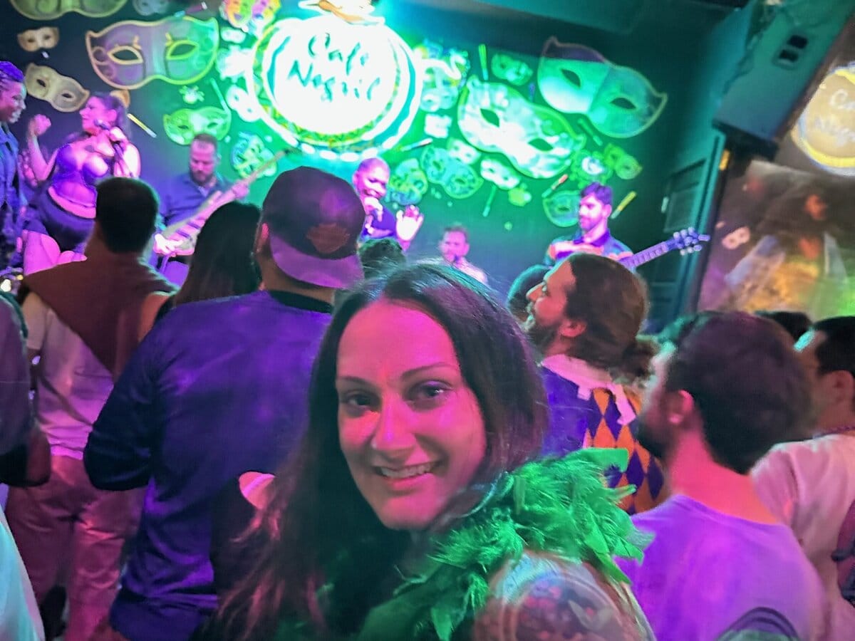 A woman wearing a green feather boa smiles at the camera in a lively, crowded bar or club with people watching a live band perform on a brightly lit stage decorated with green and purple masks.