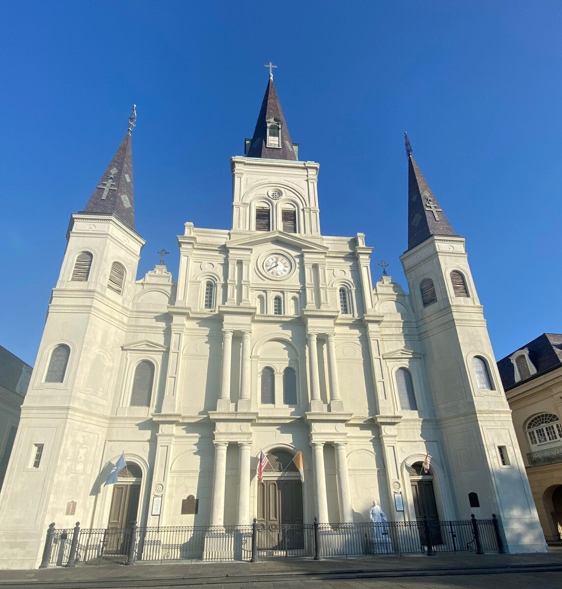 A tall, white cathedral with three steeples and a clock at the center stands against a clear blue sky—a must-see among free things to do in New Orleans. The entrance features double doors, arched windows, and flags above the entryway.