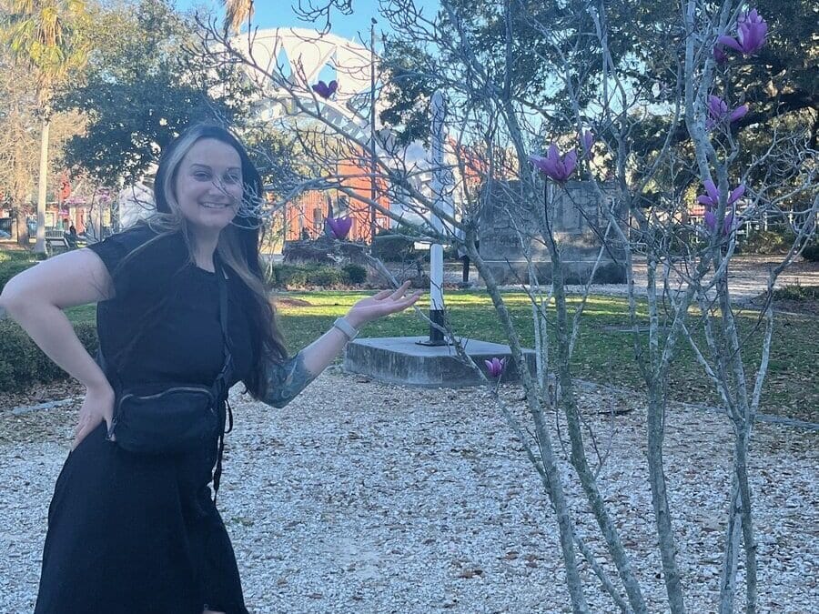 A smiling woman in a black dress stands outdoors next to a small tree with purple flowers, gesturing toward the blossoms in a park setting with trees and sunlight in the background.