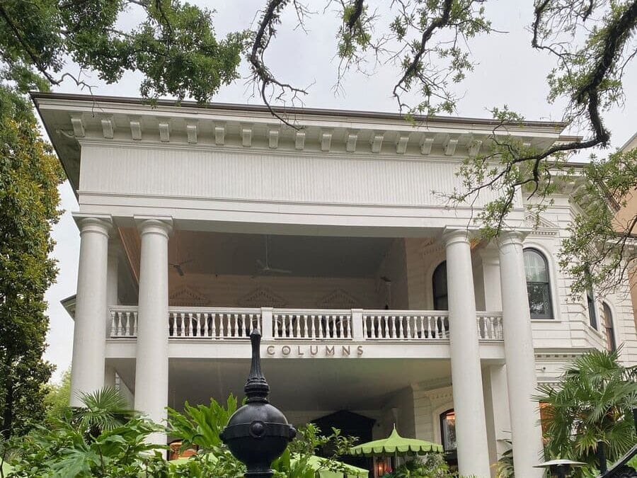 A large white historic building with tall columns, a balcony, arched windows, and lush green plants in front. The word COLUMNS is displayed above the entrance.