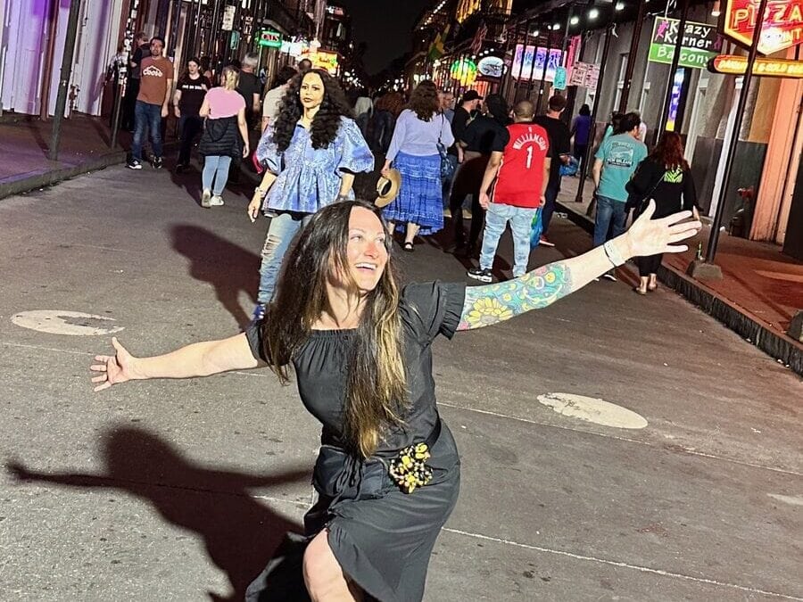 A woman with long hair and a floral tattoo poses joyfully in a black dress on a lively city street at night, while people walk and neon signs glow in the background.
