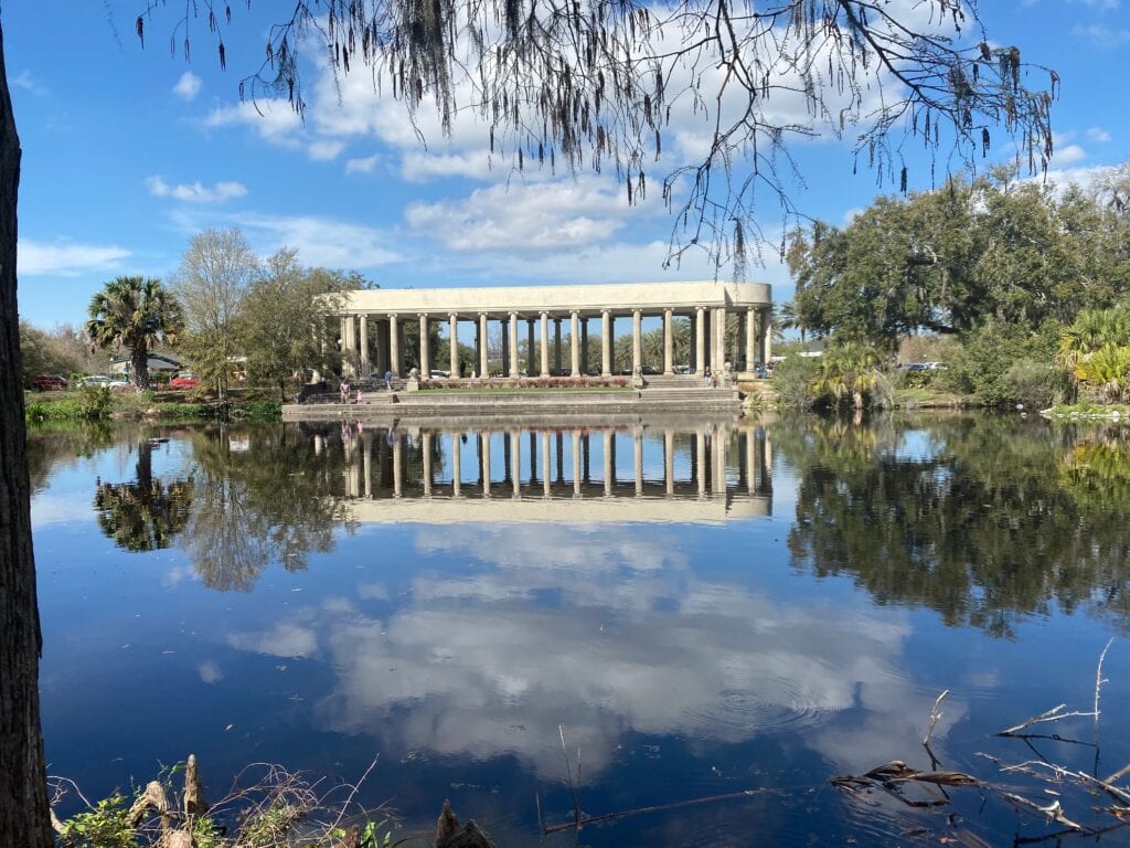 A rectangular building with columns is reflected in a calm lake, surrounded by trees and blue sky. This tranquil scene, featuring lush greenery and mirrored water, captures the beauty of free things to do in New Orleans.