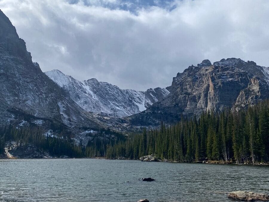 A serene mountain lake bordered by dense pine trees, with rugged, snow-capped peaks rising in the background under a partly cloudy sky.