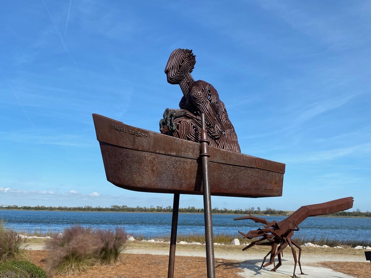 A metal sculpture of a person sitting in a boat is elevated on poles near a waterfront, with bushes and a second abstract sculpture visible in the background under a clear blue sky.