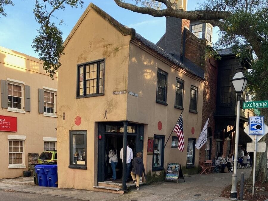A small, historic two-story building with American flags, people entering, and a faded sign on the corner of a street with crosswalk lines. Blue recycling bins sit outside, and a street sign reads “Exchange.”.