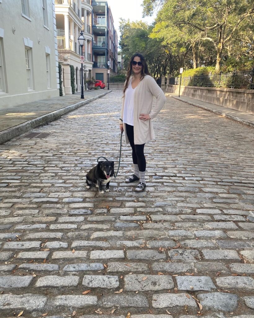 A woman wearing sunglasses and casual clothes stands on a cobblestone street holding a leash attached to a small black and white dog sitting beside her. Trees and buildings line the street in the background.