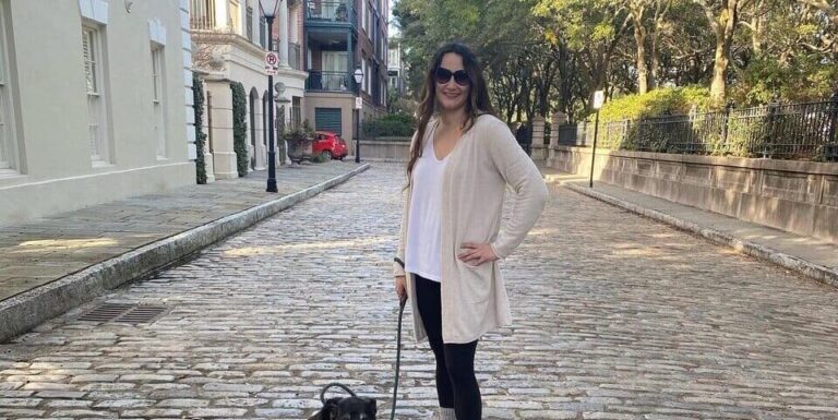 A woman wearing sunglasses and casual clothes stands on a cobblestone street holding a leash attached to a small black and white dog sitting beside her. Trees and buildings line the street in the background.
