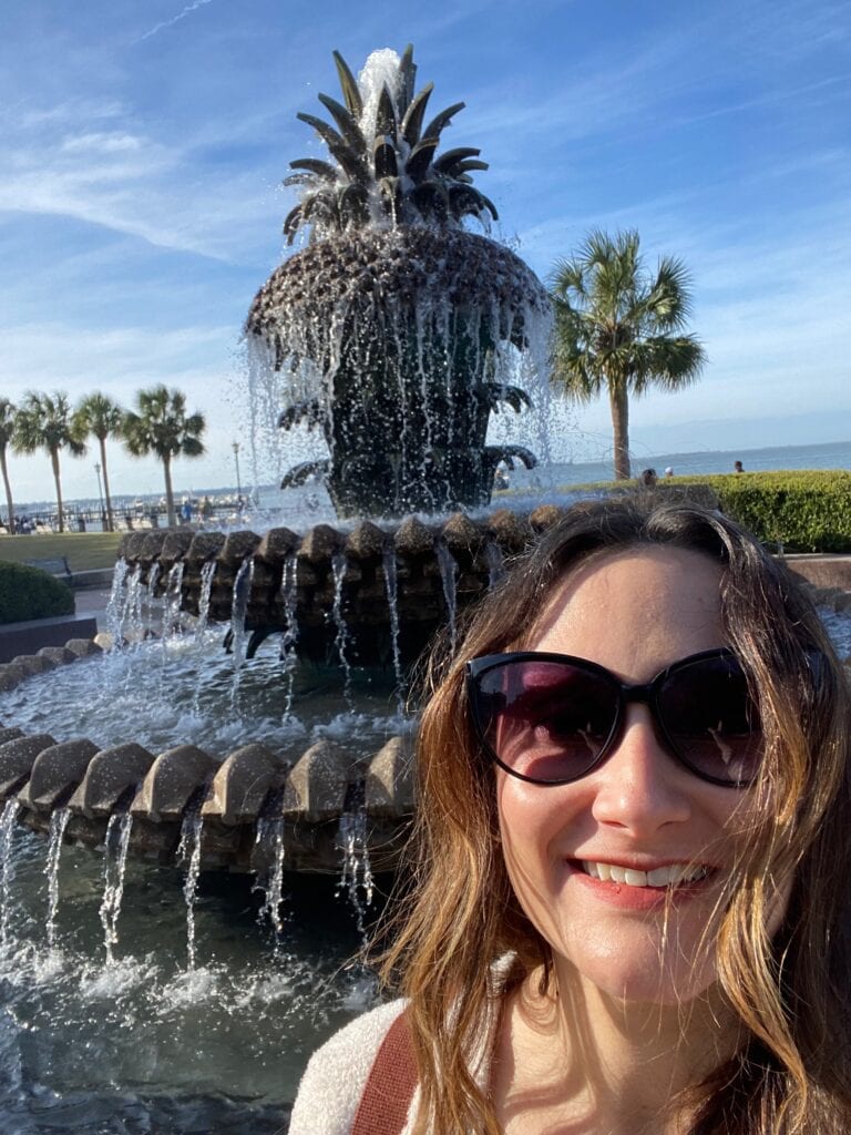 A woman wearing sunglasses smiles in front of a large pineapple-shaped fountain with water cascading down, palm trees, and a blue sky in the background.
