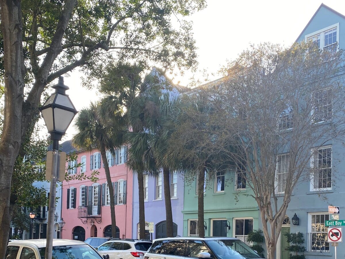 Cars are parked and driving on a city street lined with palm trees and pastel-colored buildings, including pink, blue, and green facades, on a sunny day.