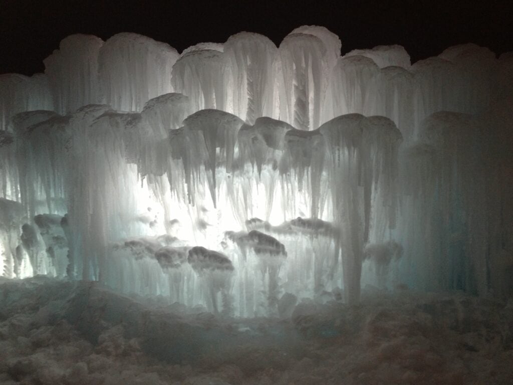 A cluster of tall, thick icicles illuminated from behind, creating a glowing, layered ice formation against a dark background. Snow blankets the ground beneath the frozen structures.