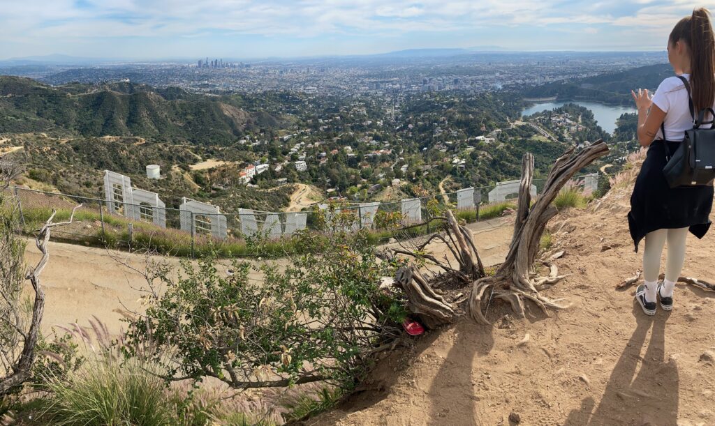 A woman stands near the Hollywood Sign overlooking Los Angeles; the city skyline, hills, and a reservoir are visible under a partly cloudy sky. Shrubs and a dirt trail appear in the foreground.