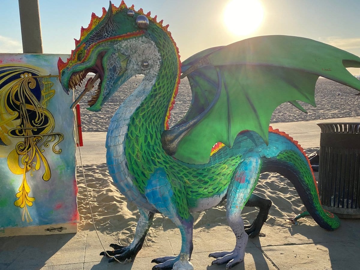 A colorful dragon sculpture with green wings stands on a sunlit beach sidewalk, casting long shadows. The sun is low in the sky, and nearby is a painted box and a trash can.