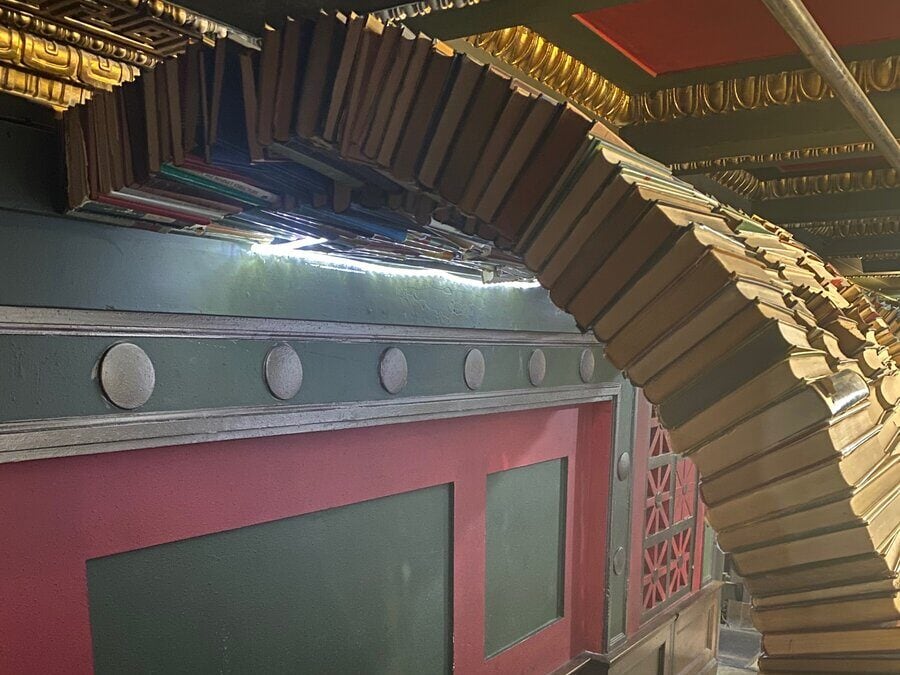 A spiraling archway made of stacked books curves over a dark hallway with red and green walls, gold accents, and circular decorative elements. Sunlight filters through a small gap above.