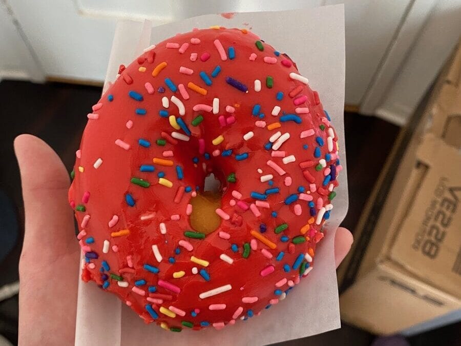 A hand holds a pink-frosted donut with colorful sprinkles on a piece of white paper. In the background, theres a cardboard box and a striped slipper on a dark floor.
