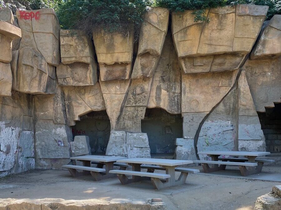 Three picnic tables are set in front of a rock wall with several cave-like openings. The rocks have some graffiti and are surrounded by greenery under a sunny sky. Shadows from nearby trees fall across the scene.