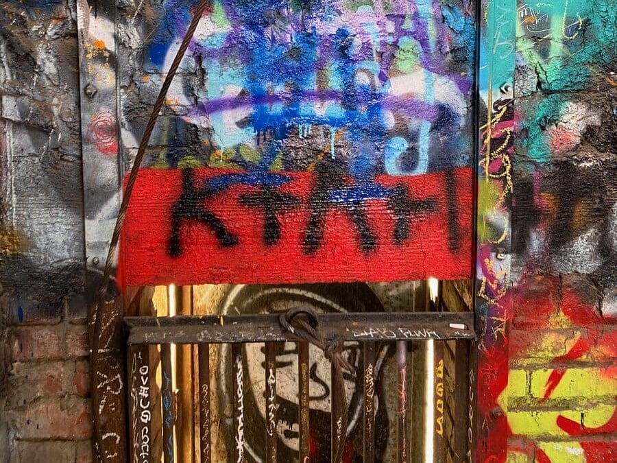 A vibrant, colorful wall covered with overlapping layers of graffiti, paint splatters, and tags. A prominent red rectangle at the bottom features bold black letters, partially obstructed by a metal gate in the foreground.