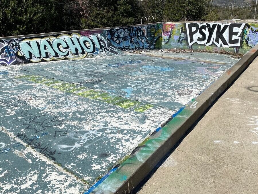 An empty, abandoned swimming pool with peeling paint and graffiti on the walls, including the words NACHO and PSYKE, surrounded by trees and hills under a clear blue sky.