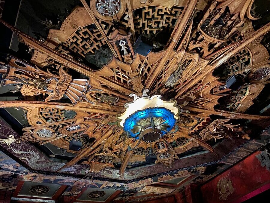Ornate, spiked ceiling centerpiece with intricate gold patterns, blue and white glowing light at the center, set in a dimly lit, richly decorated room with dark walls and red accents.