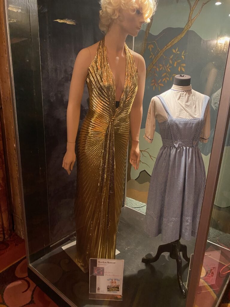 A mannequin wears a gold, pleated, halter-neck gown next to another mannequin dressed in a blue-and-white gingham dress with a white blouse, both displayed in a glass case with a small information plaque in front.