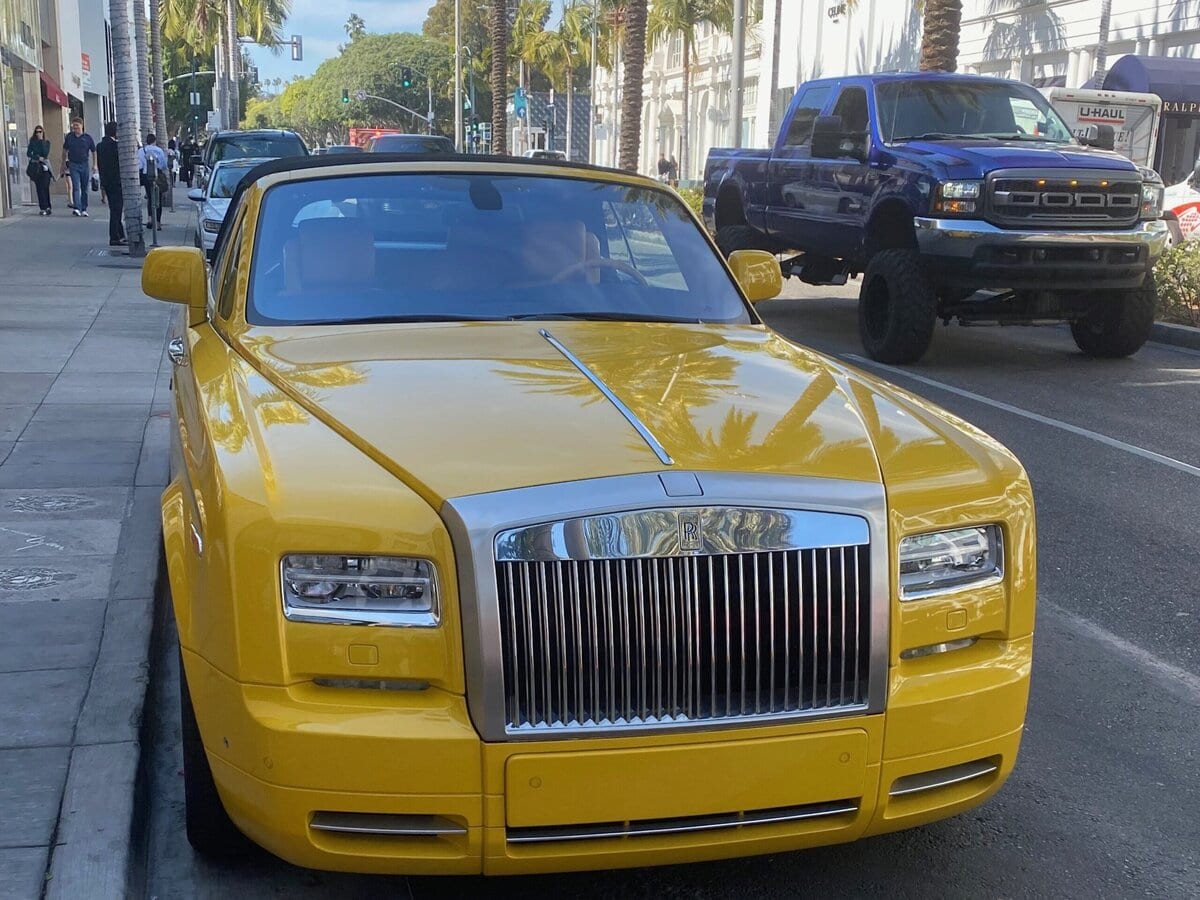 A bright yellow luxury convertible car is parked by the sidewalk on a palm tree-lined street, with a large black pickup truck and other vehicles in the background.
