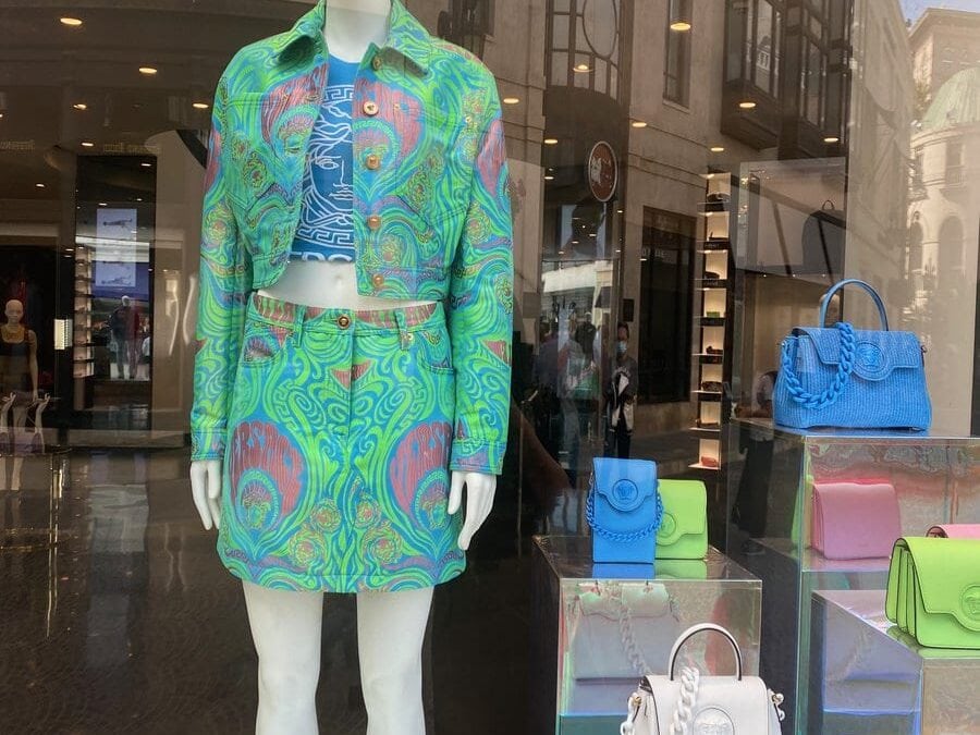 A mannequin in a store window displays a vibrant green and blue patterned jacket and skirt set with a matching top. Nearby, colorful blue, green, and white handbags are arranged on display shelves.