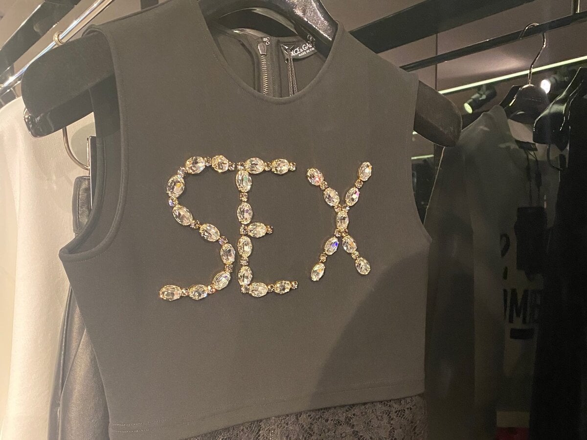 A sleeveless black crop top with lace trim displayed on a hanger, featuring the word SEX spelled out in large, shiny rhinestones across the chest.