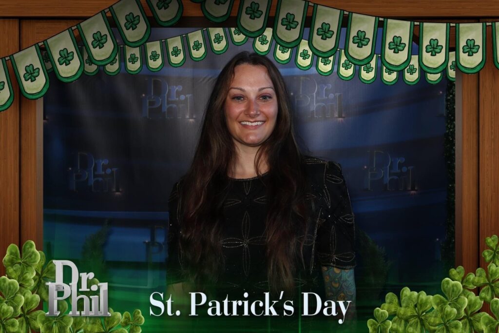 A smiling woman with long dark hair stands in front of a Dr. Phil backdrop, surrounded by St. Patrick’s Day decorations with shamrocks and a banner. St. Patrick’s Day is written at the bottom of the image.