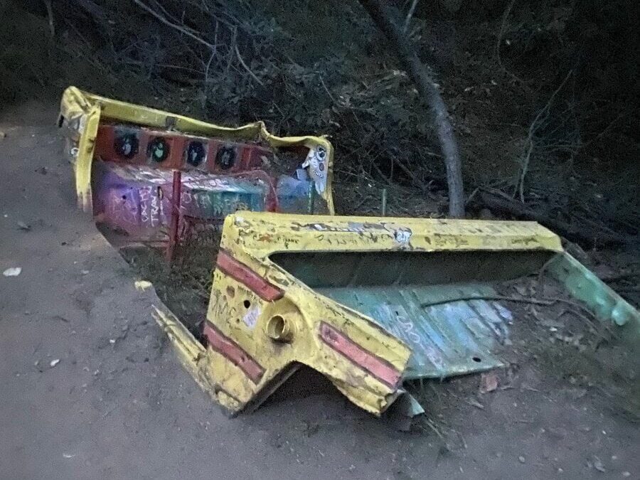 Two large, colorful, rusted metal pieces, possibly from an amusement park ride, lie abandoned on a dirt path, surrounded by trees and foliage in a dimly lit, outdoor setting.