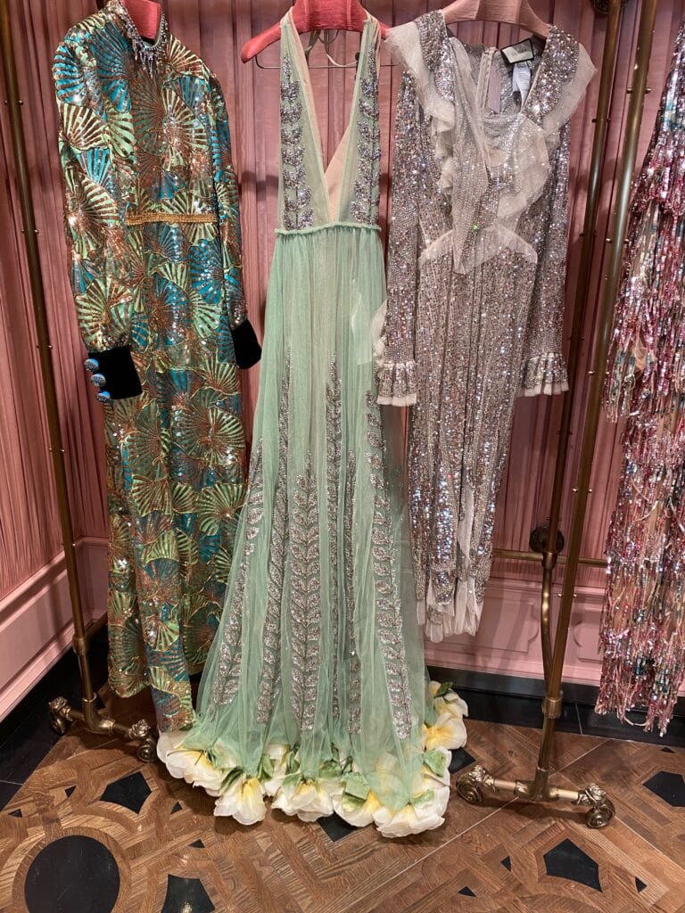 Three ornate dresses on display: a high-neck dress with green, gold, and blue geometric patterns; a sleeveless, sheer mint gown with floral accents; and a long-sleeve silver sequin dress with ruffles, all against a pink backdrop.