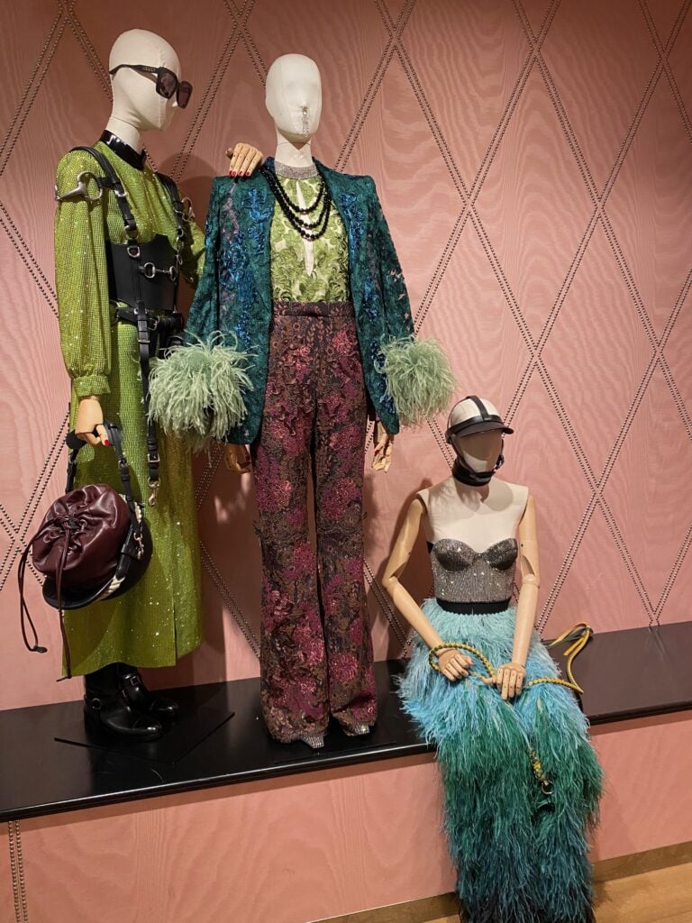 Three mannequins display vibrant outfits: one in a green dress with harness and sunglasses, one in a green and teal feather-trimmed suit with patterned pants, and one seated in a feathered teal skirt and silver beaded bra top.