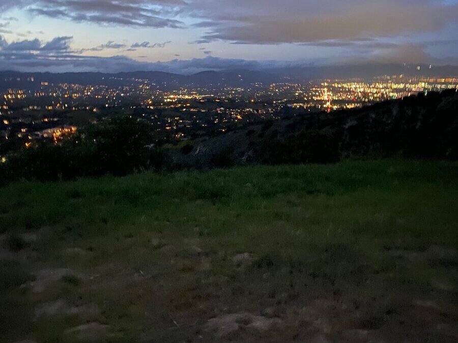 A grassy hilltop at dusk overlooks a cityscape filled with glowing lights. The sky is partly cloudy, with fading daylight and a soft blue hue above the landscape.