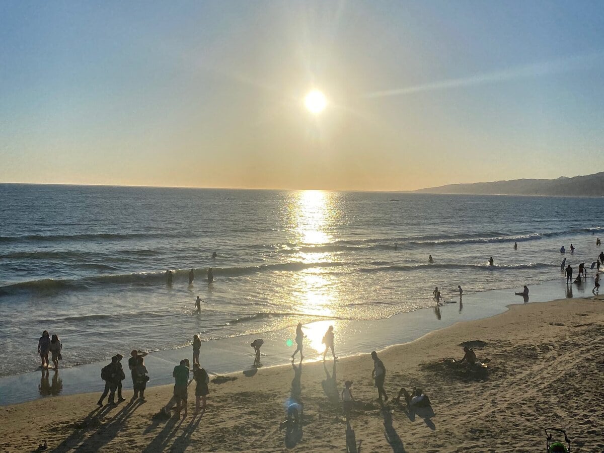 People enjoy a sunny day at the beach, with the sun low in the sky, casting long shadows on the sand while others swim and play in the ocean. The sea reflects the sunlight, creating a bright path on the water.