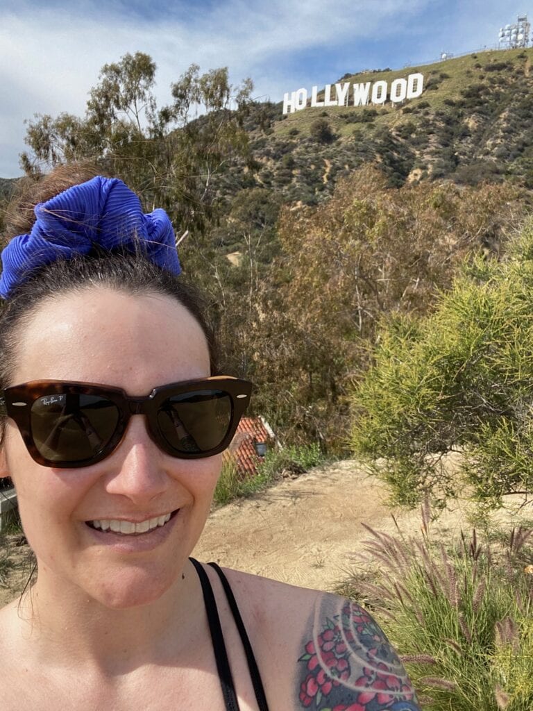 A woman wearing sunglasses and a blue scrunchie smiles in front of the Hollywood sign on a sunny day, with trees and shrubs around her and hills in the background.