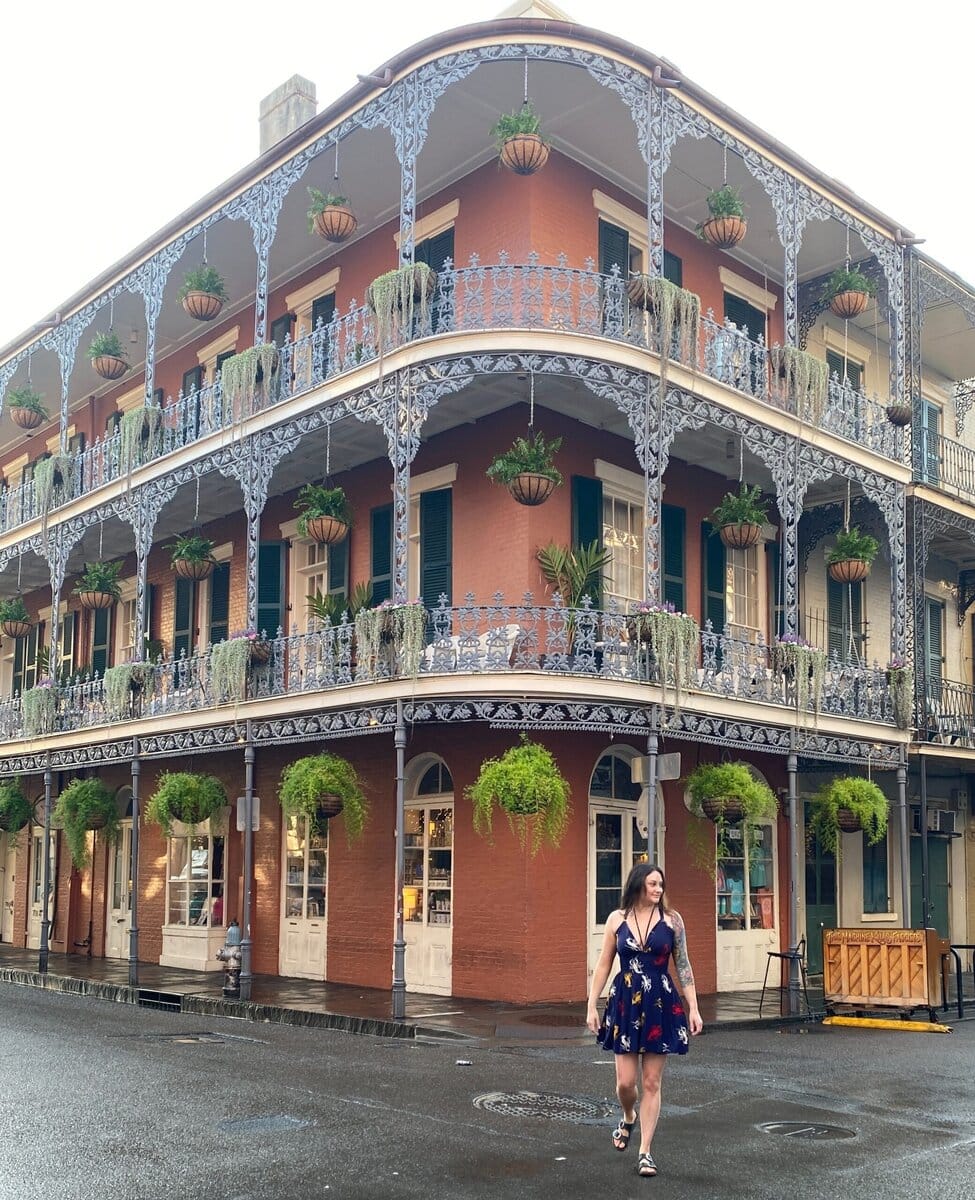 A woman in a blue dress walks across a wet street in front of a historic brick building with ornate ironwork balconies and hanging plants in New Orleans.
