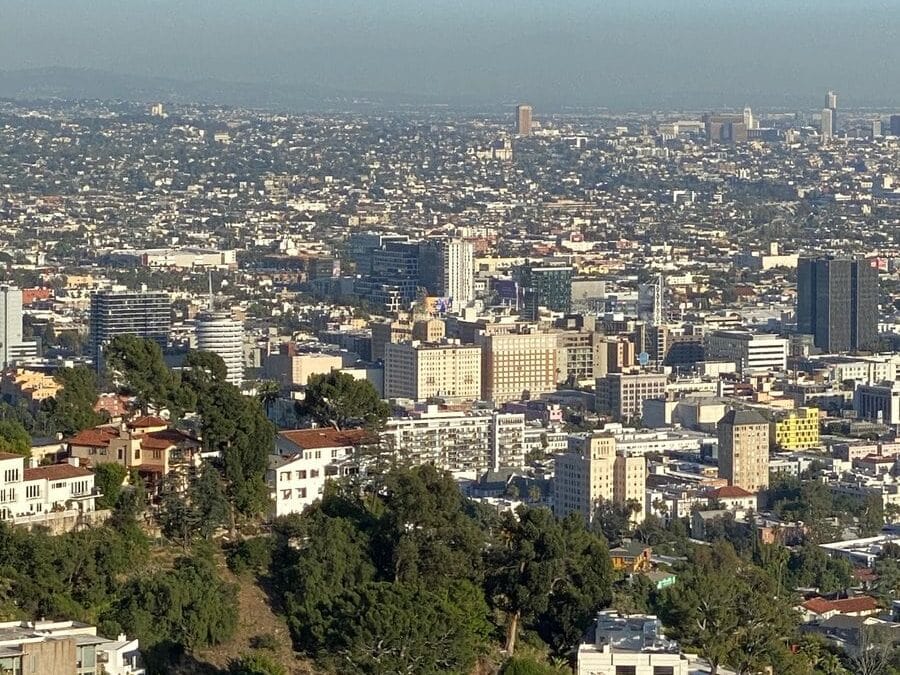 A wide view of a cityscape with numerous buildings, trees, and hills in the foreground under a clear blue sky. The area appears densely populated, stretching far into the distance.